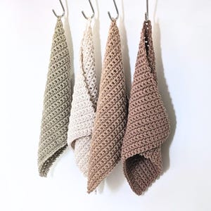 May include: Four square crochet washcloths hanging from silver metal hooks. The cloths are textured with a dense stitch pattern and come in a gradient of neutral earth tones including sage green, cream, light tan, and a medium brown shade.