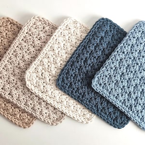 May include: Five square crochet washcloths arranged in a row, featuring a textured shell stitch pattern. The colours include light tan, beige, cream, dark navy blue, and light blue, all crafted from thick yarn with visible interlocking loops.