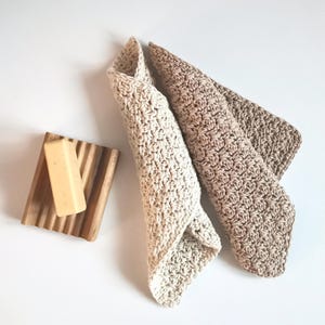 May include: Two folded, cream-colored crochet washcloths and one tan crochet washcloth are displayed on a white surface. A wooden soap dish with a bar of soap is also present. The washcloths have a textured appearance.