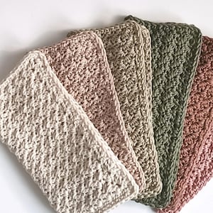Può includere: Una collezione di cinque asciugamani all'uncinetto fatti a mano in una varietà di colori. Gli asciugamani sono disposti a ventaglio, mostrando le diverse tonalità di crema, rosa, verde oliva e malva. Il motivo a punto strutturato è visibile.