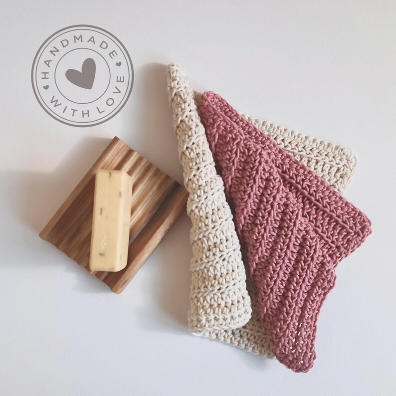May include: Handmade with Love. A wooden soap dish with a bar of soap and two crocheted washcloths in cream and dusty rose. The washcloths are folded and arranged on a white surface.