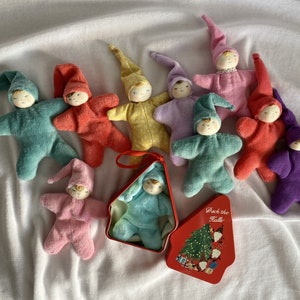 Puede incluir: Una colección de muñecas elfo hechas a mano en varios colores, incluyendo turquesa, rojo, amarillo, morado y rosa. Cada muñeca tiene un cuerpo en forma de estrella, un gorro puntiagudo y una cara pintada. Una muñeca está en una lata roja con un diseño de árbol de Navidad.