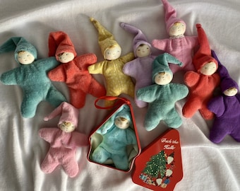 Può includere: Una collezione di bambole elfo fatte a mano in vari colori, tra cui verde acqua, rosso, giallo, viola e rosa. Ogni bambola ha un corpo a forma di stella, un cappello a punta e un viso dipinto. Una bambola è in una scatola di latta rossa con un disegno di albero di Natale.