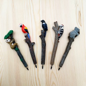 Peut inclure: Une collection de cinq stylos décoratifs avec des figurines d'animaux. Les stylos comprennent un paresseux, un perroquet, un corbeau, un pic et un koala. Chaque stylo a une figurine d'animal détaillée perchée sur une fausse branche en bois.