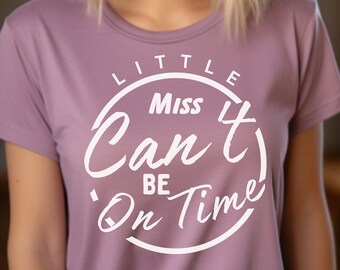 May include: A mauve-colored t-shirt with the text "Little Miss Can't Be On Time" in a white, circular design. The shirt is a crew neck style and appears to be made of a soft material. The image is of a person wearing the shirt.