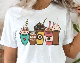 Pode incluir: Camiseta branca com um gráfico colorido de seis bebidas de café diferentes. Cada bebida tem coberturas e cores únicas, incluindo chantilly, chocolate e frutas. O texto "Dog Mom" também está incluído.