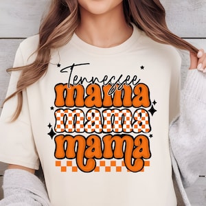 May include: Cream-colored t-shirt with the text "Tennessee Mama" in orange and black. The design includes a checkered pattern and star accents. The shirt is a casual, comfortable style.