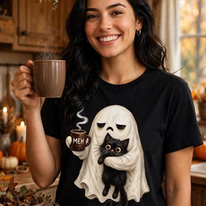May include: A black short-sleeved t-shirt featuring a graphic of a white ghost holding a steaming mug labeled MEH and hugging a black cat. The ghost has a sad expression, and the cat has large yellow eyes. The design is centered on the chest.