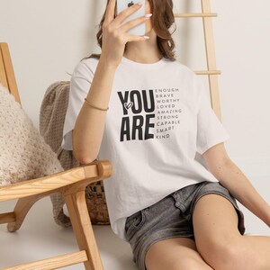 Könnte beinhalten: Ein weißes kurzärmeliges T-Shirt mit dem schwarzen Text "YOU ARE", gefolgt von einer Liste der Wörter: "ENOUGH", "BRAVE", "WORTHY", "LOVED", "AMAZING", "STRONG", "CAPABLE", "SMART", "KIND", mit einem kleinen Herzsymbol, das in das Design integriert ist.