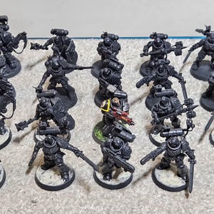 May include: A group of miniature figurines depicting armored soldiers, each equipped with weapons and equipment. The figures are predominantly dark grey, with some featuring red and yellow details. They are set on circular bases.