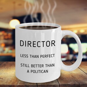 May include: White ceramic coffee mug with the text "DIRECTOR LESS THAN PERFECT STILL BETTER THAN A POLITICIAN" in black. The mug is filled with dark coffee, and steam rises from the top. The mug sits on a wooden surface.