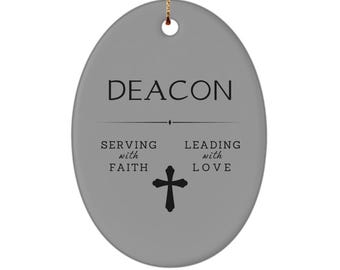May include: Oval-shaped gray ceramic ornament with the word "DEACON" in black. Below, it reads "SERVING with FAITH" and "LEADING with LOVE" with a black cross in the center. A gold string is attached for hanging.