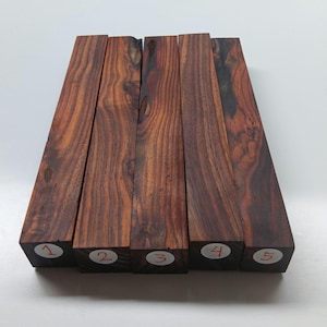 May include: Five rectangular blocks of dark brown wood with visible grain patterns. Each block has a sticker with a number from one to five. The wood has a reddish-brown hue and a polished appearance, suggesting it is for woodworking.
