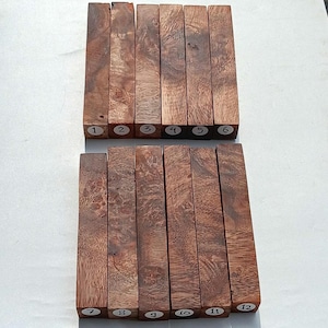 May include: Twelve rectangular wooden turning blanks arranged in two rows of six. Each piece features a distinct brown wood grain pattern with natural swirls and knots. Every blank is labeled with a small white circular sticker numbered one through twelve.