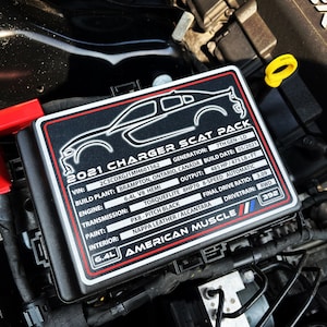 May include: Close-up of a black engine compartment label with white text. The label reads "2021 Charger Scat Pack" and includes details about the vehicle's engine, transmission, and other specifications. The label also features the text "American Muscle" in red and white.
