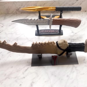 Może przedstawiać: Trzy dekoracyjne noże na wystawie. Górny nóż ma złote ostrze i czarny uchwyt. Środkowy nóż ma srebrne ostrze i brązowy uchwyt. Dolny nóż jest w kolorze kości z czarnym uchwytem i paskiem. Widoczny jest tekst "CARRY ON MY WAYWARD SON".