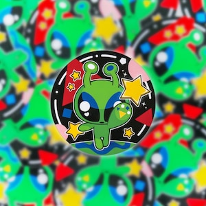 May include: A circular sticker with a cartoon green alien character with large blue eyes, waving. The design includes yellow stars and geometric shapes on a black background. The background has a repeating pattern of aliens and stars.
