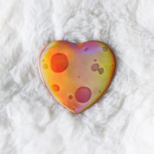 May include: A heart-shaped object with a colorful, iridescent surface. The colors transition from orange to yellow and purple, with several circular, darker spots. The object is set against a white, fluffy background.