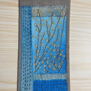 May include: Embroidered textile art with a blue and grey floral design. The rectangular piece has a grey border and a hanging loop. The central design features embroidered branches with small yellow flowers, showcasing a handcrafted aesthetic.