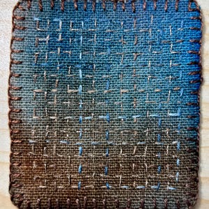 May include: A square fabric patch featuring a gradient colour transition from blue at the top to brown at the bottom. The textile is decorated with a grid of hand-stitched embroidery in light blue and tan thread, finished with a brown blanket stitch.