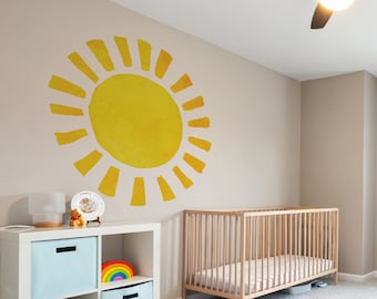 May include: A white crib with a wooden frame and a white mattress sits in a nursery with a light gray carpet. A white bookshelf with blue and rainbow storage bins sits next to the crib. A yellow sun decal is on the wall behind the crib.