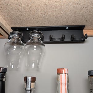 May include: A black, under-cabinet wine glass rack with two clear wine glasses hanging from it. The rack is mounted under a wooden shelf. Several bottles with various caps are visible below the glasses.