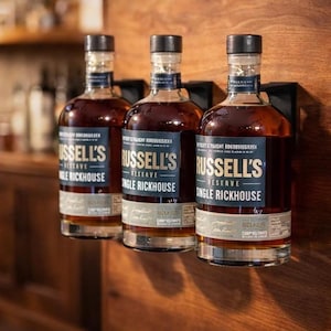 May include: Three bottles of Russell's Reserve Single Rickhouse bourbon are displayed on a wooden surface. The bottles have dark glass with black caps and labels with white text. The bottles are held in place by black metal brackets.