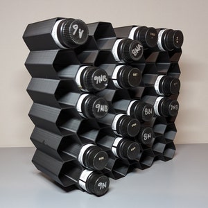 May include: A black, honeycomb-shaped bottle rack holding multiple bottles. The bottles have black caps with white labels and various handwritten markings. The rack is made of a dark material and is designed to hold the bottles at an angle.