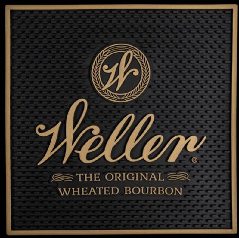 Pu&ograve; includere: Tappetino da bar nero con superficie strutturata e bordo dorato. Il tappetino presenta il nome "Weller" in elegante scrittura, un emblema circolare e il testo "THE ORIGINAL WHEATED BOURBON."