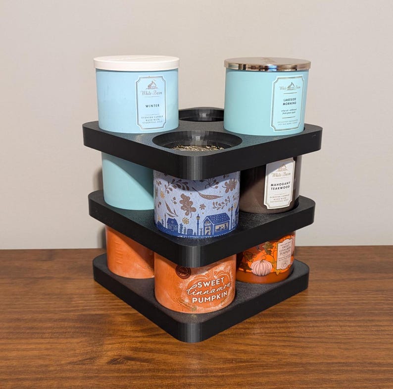 K&ouml;nnte beinhalten: Ein schwarzer, dreist&ouml;ckiger Kerzenhalter mit verschiedenen Duftkerzen. Die Kerzen sind in Blau-, Orange- und Braunt&ouml;nen gehalten, mit Etiketten, die D&uuml;fte wie "Winter" und "Sweet Cinnamon Pumpkin" angeben. Der Halter besteht aus schwarzem Material.