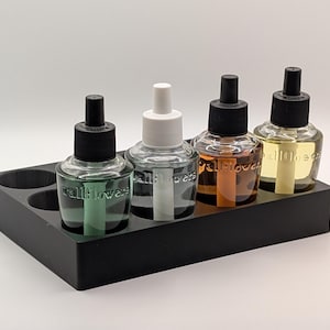 May include: Four glass air freshener refills with black caps and various colored liquids, displayed in a black rectangular holder. The refills are labeled with the word "wallflowers".
