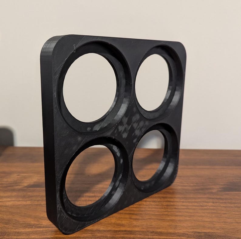 K&ouml;nnte beinhalten: Ein schwarzes, quadratisches Objekt mit vier kreisf&ouml;rmigen &Ouml;ffnungen. Das Objekt scheint 3D-gedruckt zu sein und hat eine strukturierte Oberfl&auml;che. Die &Ouml;ffnungen sind gleichm&auml;&szlig;ig verteilt und von unterschiedlicher Gr&ouml;&szlig;e. Es liegt auf einer Holzoberfl&auml;che.