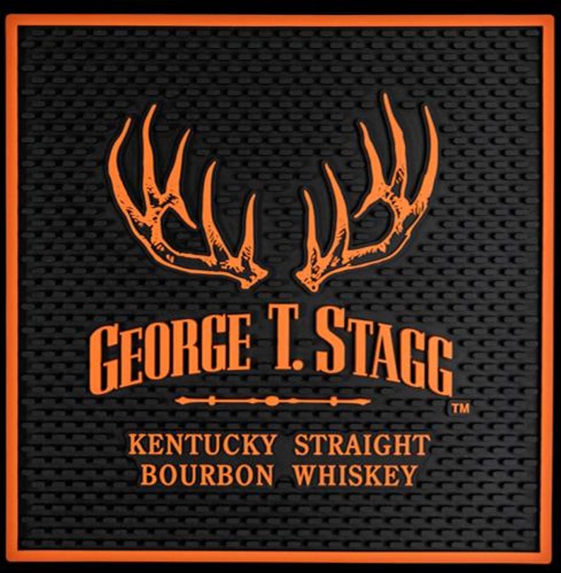 Pu&ograve; includere: Tappetino da bar nero con bordo arancione e motivo ripetuto. Il tappetino presenta il testo "GEORGE T. STAGG" e "KENTUCKY STRAIGHT BOURBON WHISKEY" in arancione, insieme a un design a corna.