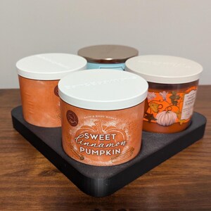 K&ouml;nnte beinhalten: Vier Bath & Body Works-Kerzen mit wei&szlig;en Deckeln stehen auf einem schwarzen Tablett. Auf der vorderen Kerze steht "Sweet Cinnamon Pumpkin" mit einer K&uuml;rbisgrafik. Die anderen Kerzen sind orange und hellblau.