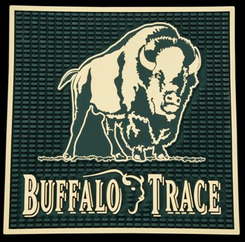 Pu&ograve; includere: Un tappetino da bar verde scuro con una superficie strutturata e un contorno color crema. Il tappetino presenta un'illustrazione color crema di un bisonte e le parole "BUFFALO TRACE" in un font stilizzato.