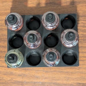 K&ouml;nnte beinhalten: Eine schwarze Silikonform mit neun runden Vertiefungen, jede mit einer kleinen Glasflasche mit schwarzem Deckel. Die Flaschen sind mit einer klaren Fl&uuml;ssigkeit gef&uuml;llt.