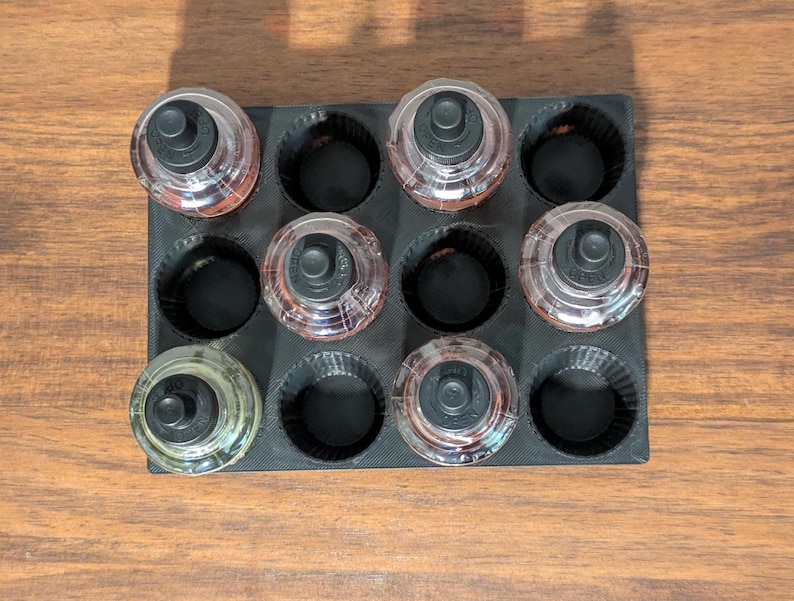 K&ouml;nnte beinhalten: Eine schwarze Silikonform mit neun runden Vertiefungen, jede mit einer kleinen Glasflasche mit schwarzem Deckel. Die Flaschen sind mit einer klaren Fl&uuml;ssigkeit gef&uuml;llt.
