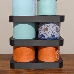 K&ouml;nnte beinhalten: Ein schwarzer, mehrst&ouml;ckiger Kerzenhalter mit vier Duftkerzen. Die Kerzen sind in verschiedenen Farben gehalten, darunter Hellblau, Orange und ein Blumenmuster. Der Halter besteht aus dunklem Material und steht auf einer Holzoberfl&auml;che.