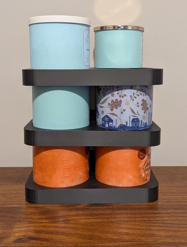 K&ouml;nnte beinhalten: Ein schwarzer, mehrst&ouml;ckiger Kerzenhalter mit vier Duftkerzen. Die Kerzen sind in verschiedenen Farben gehalten, darunter Hellblau, Orange und ein Blumenmuster. Der Halter besteht aus dunklem Material und steht auf einer Holzoberfl&auml;che.