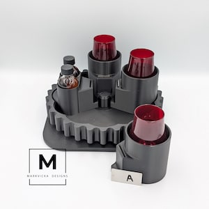 May include: A black, gear-shaped bar caddy with four red-tinted glasses and two small bottles. The caddy has a central rotating mechanism and a label that reads "A". The logo "M" and "MARKVICKA DESIGNS" are also visible.
