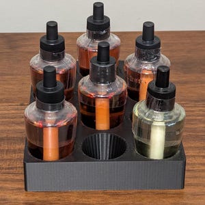 K&ouml;nnte beinhalten: Ein schwarzer Plastikhalter mit sechs durchsichtigen Glasflaschen mit schwarzen Kappen und Tropfern. Die Flaschen sind mit verschiedenen Fl&uuml;ssigkeiten gef&uuml;llt, darunter braun, orange und klar.