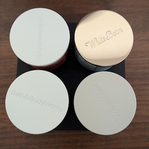 K&ouml;nnte beinhalten: Vier runde Beh&auml;lter mit Deckeln. Zwei Deckel sind wei&szlig; mit gepr&auml;gtem "Bath & Body Works", und zwei sind gold und wei&szlig; mit gepr&auml;gtem "White Barn". Die Beh&auml;lter stehen auf einem schwarzen Quadrat auf einer Holzoberfl&auml;che.