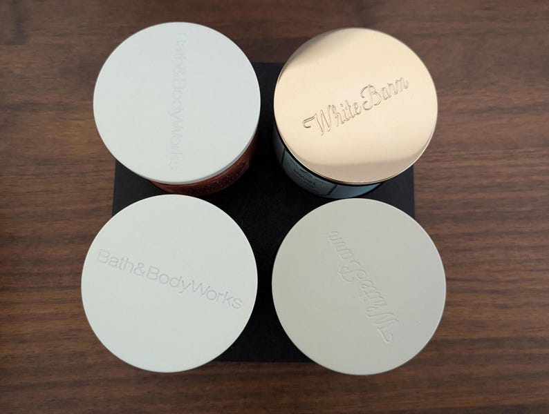 K&ouml;nnte beinhalten: Vier runde Beh&auml;lter mit Deckeln. Zwei Deckel sind wei&szlig; mit gepr&auml;gtem "Bath & Body Works", und zwei sind gold und wei&szlig; mit gepr&auml;gtem "White Barn". Die Beh&auml;lter stehen auf einem schwarzen Quadrat auf einer Holzoberfl&auml;che.
