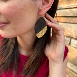 May include: Black and gold leather earrings with a teardrop shape. The earrings are hanging from a person's ear.