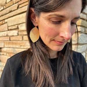 May include: A woman wearing a black shirt with gold leather earrings. The earrings are shaped like a leaf and have a textured surface.