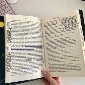 Puede incluir: Un libro abierto con texto resaltado y notas, sostenido por una persona que lleva pulseras. La portada del libro es negra con detalles dorados. Las páginas están llenas de texto.