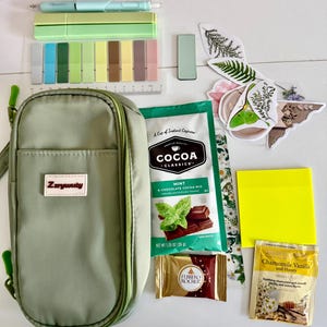 May include: An assortment of stationery and snacks, including a green pencil case, pens, sticky notes, and a packet of mint and chocolate cocoa mix. Also includes a Ferrero Rocher chocolate and chamomile vanilla tea.