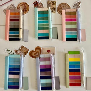 May include: A collection of colorful pens and color swatches arranged on a white surface. Each pen has a matching set of color swatches. Decorative stickers and a keyboard are also visible in the background. The pens and swatches are in various pastel colors.