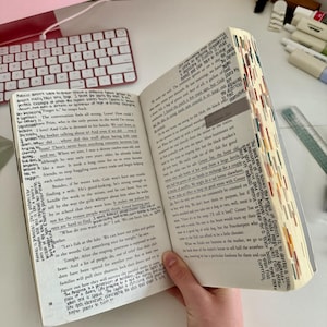 May include: An open book with multi-coloured page markers, held in a hand wearing a gold bracelet. Text is visible, with words like "Prim" and "Gale". A pink keyboard and other items are in the background.