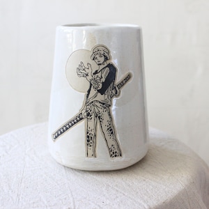 Könnte beinhalten: Eine weiße Keramikvase mit einer schwarz-weißen Illustration einer Figur, die ein Schwert hält. Die Illustration zeigt eine Person mit Hut, Hemd und gemusterter Hose. Die Vase hat eine leicht konische Form.